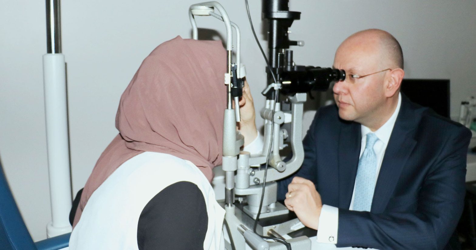 Woman wearing a hijab looks into an eye testing machine facing a doctor looking into the scope from the other side.